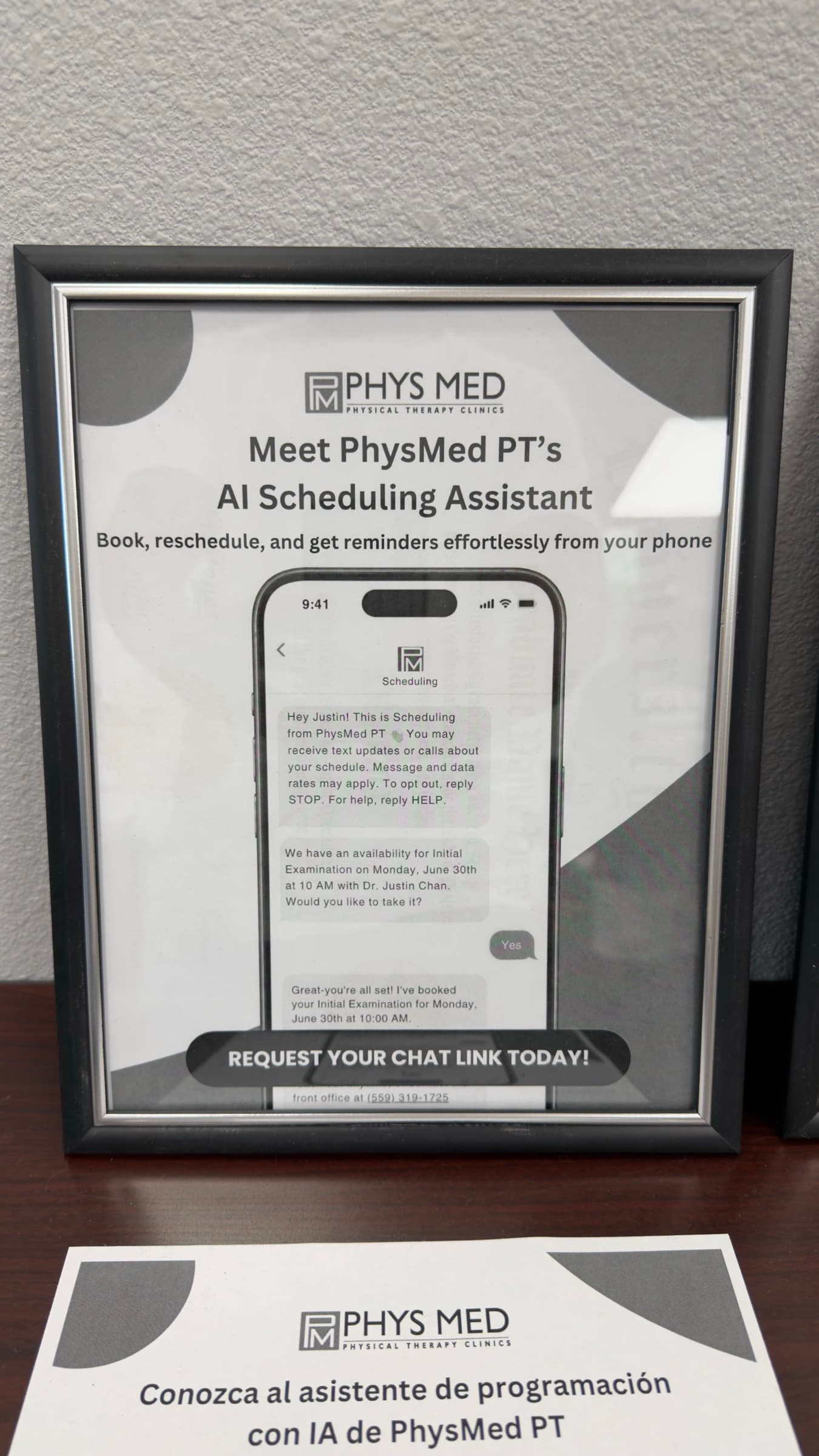 Framed poster at the Phys Med front desk introducing the clinic's AI scheduling assistant, with a Spanish-language version alongside